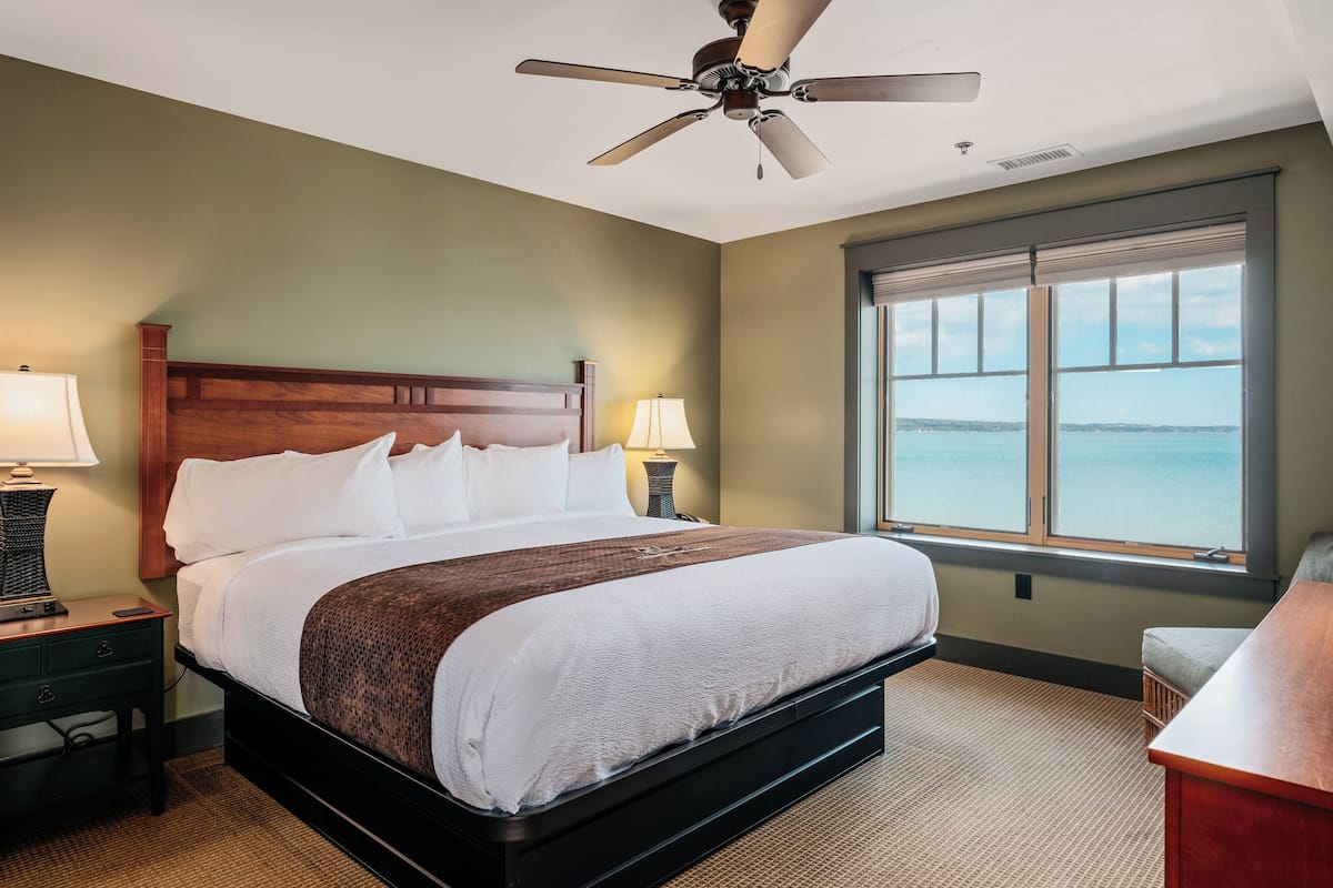 A hotel room with a large bed, two bedside lamps, a ceiling fan, and a view of the sea through a large window.