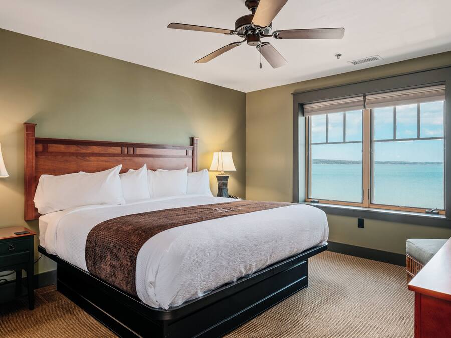 Une chambre d’hôtel avec un grand lit, deux lampes de chevet, un ventilateur de plafond et une vue sur la mer par une grande fenêtre.