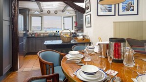 A dining area with a wooden table set for four, a view of the kitchen, and a window overlooking the sea. - 3 Bed - Smugglers Cottage (Torquay)