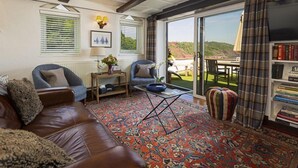 A cozy living room with a leather sofa, a blue armchair, a patterned rug, and a bookshelf. - 3 Bed - Smugglers Cottage (Torquay)