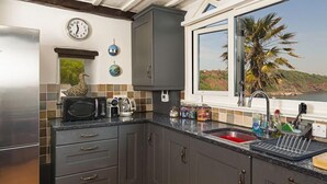 A kitchen with grey cabinets, a stainless steel refrigerator, a microwave, and a window with a view of palm trees and hills. - 3 Bed - Smugglers Cottage (Torquay)