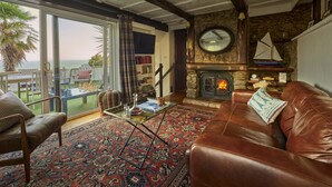 A cozy living room with a stone fireplace, leather sofa, and a view of the sea through large glass doors. - 3 Bed - Smugglers Cottage (Torquay)