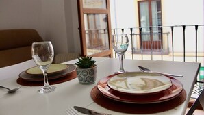 Une table à manger dressée avec des assiettes, des couverts et des verres, agrémentée d’une petite plante en pot et offrant une vue sur un bâtiment par la fenêtre.