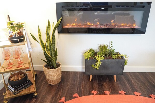 A living room with a fireplace, a plant shelf, a plant in a woven basket, and a bookcase with wine glasses and books.