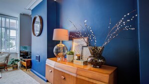 A room with a blue accent wall, a wooden dresser, a lamp, a vase with branches, and a framed picture. - Cosy house on the seafront - Pass the Keys (Southend-on-Sea)