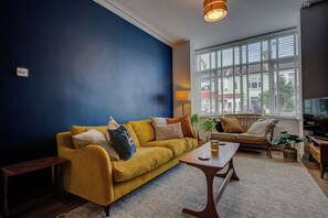 A living room with a yellow sofa, a wooden coffee table, a lamp, and a large window with blinds.