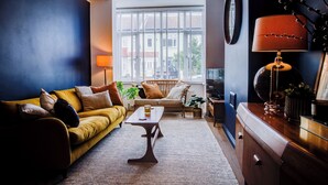 A living room with a yellow sofa, a wooden coffee table, and a large window with blinds. - Cosy house on the seafront - Pass the Keys (Southend-on-Sea)