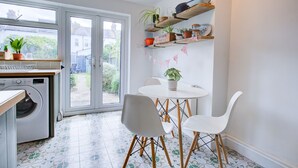 A kitchen with a washing machine, a dining table with chairs, and shelves with plants and kitchenware. - Cosy house on the seafront - Pass the Keys (Southend-on-Sea)