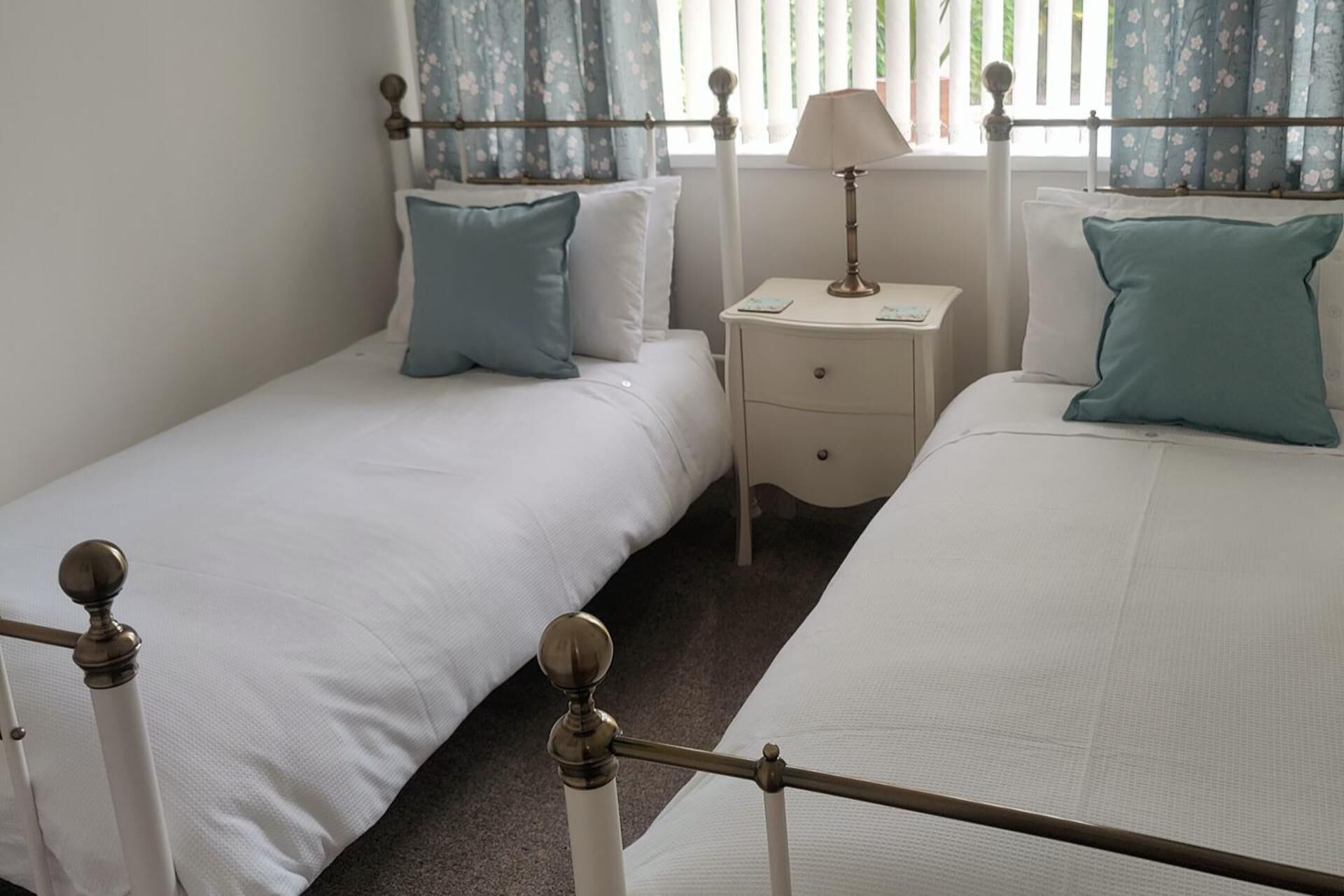 Une chambre double avec deux lits simples, une table de chevet avec une lampe et une fenêtre agrémentée de rideaux à motifs floraux.