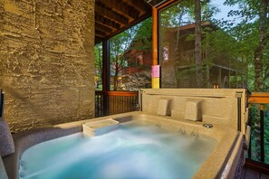 A hot tub with a stone wall and wooden deck, surrounded by trees and a wooden cabin in the background. - 3 Min to Parkway | Private Resort Pool | Top 5%! (Pigeon Forge)