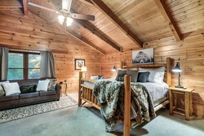 A cozy log cabin bedroom with a wooden ceiling, a bed with a wooden headboard, a nightstand, a lamp, a sofa, and a window with blinds. - 3 Min to Parkway | Private Resort Pool | Top 5%! (Pigeon Forge)