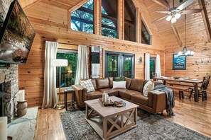 A cozy cabin interior with a wooden interior, a large window, a fireplace, a dining area, and a ceiling fan.