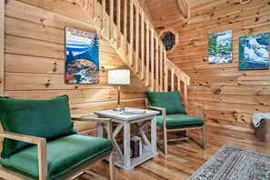 A cabin interior with wooden walls, a staircase, two green upholstered chairs, a small table with a lamp, and posters of Great Smoky Mountains National Park.