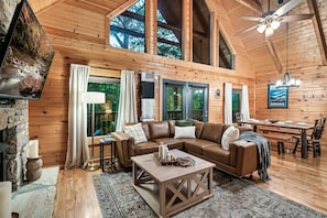 A cozy cabin interior with a wooden floor, a leather sofa, a dining table, and a large window overlooking a forest.