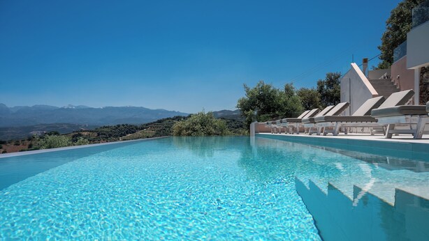 Une piscine à débordement à l’eau d’un bleu cristallin, entourée de chaises longues et offrant une vue sur des montagnes lointaines.