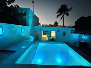 Un bâtiment moderne doté d’une piscine, d’une entrée avec une porte en verre et d’un palmier en arrière-plan.