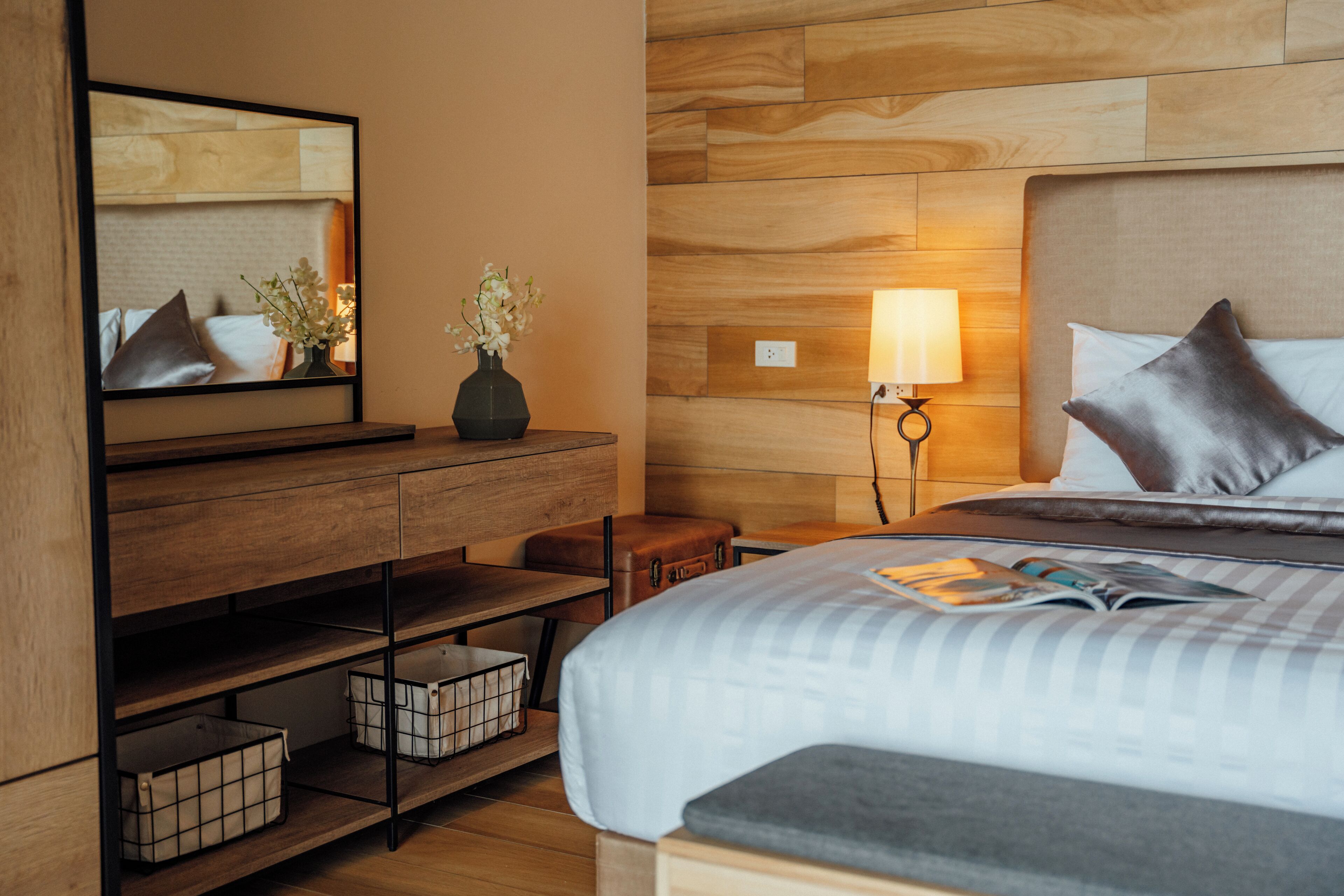 A hotel room with a wooden headboard, a bedside table with a lamp, a mirror, a dresser with storage bins, and a bed with pillows and a magazine.