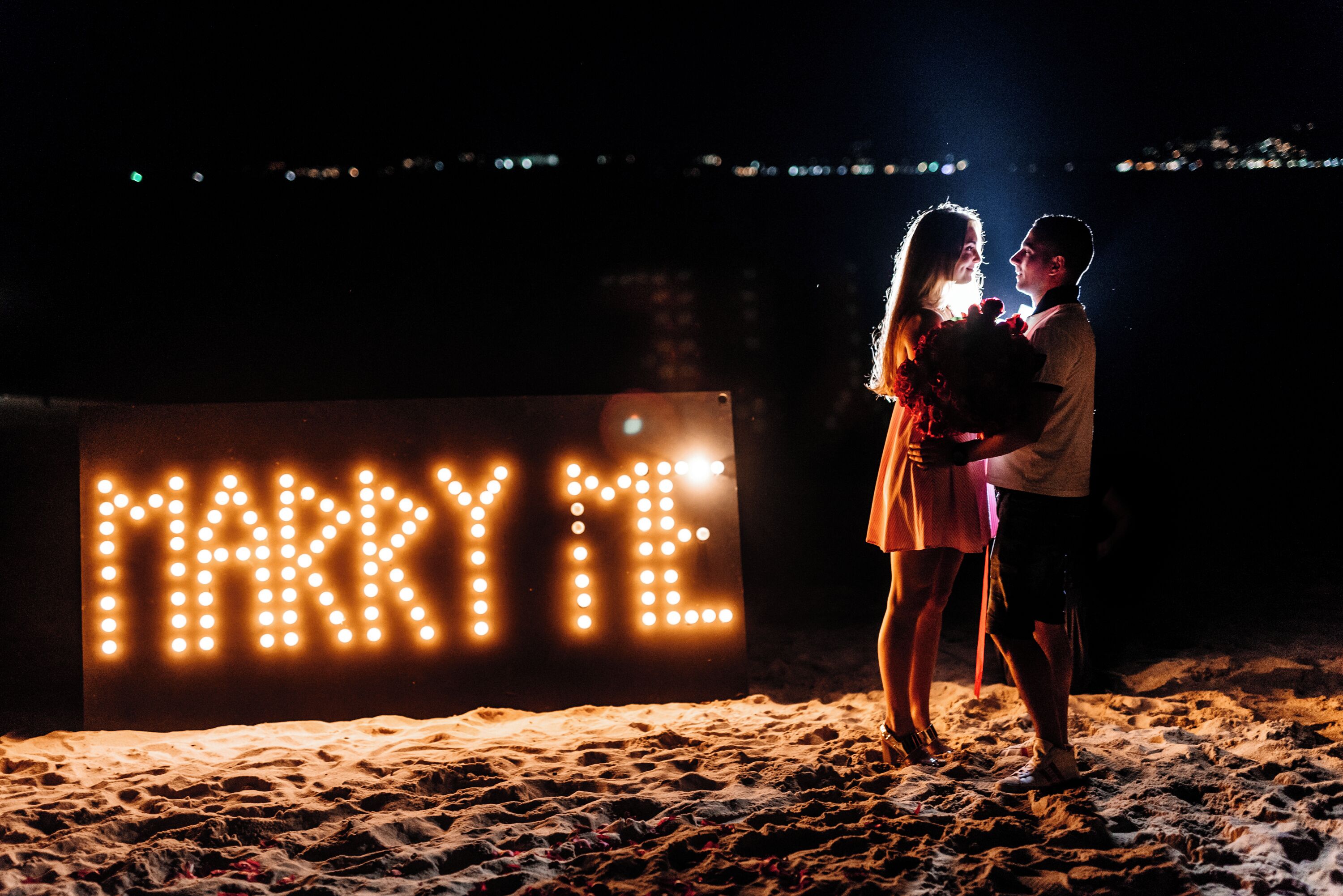 A couple on a beach at night with a 'MARRY ME' sign in the background.