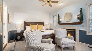 A bedroom with a bed, two armchairs, a fireplace, a ceiling fan, and a mirror on the wall.