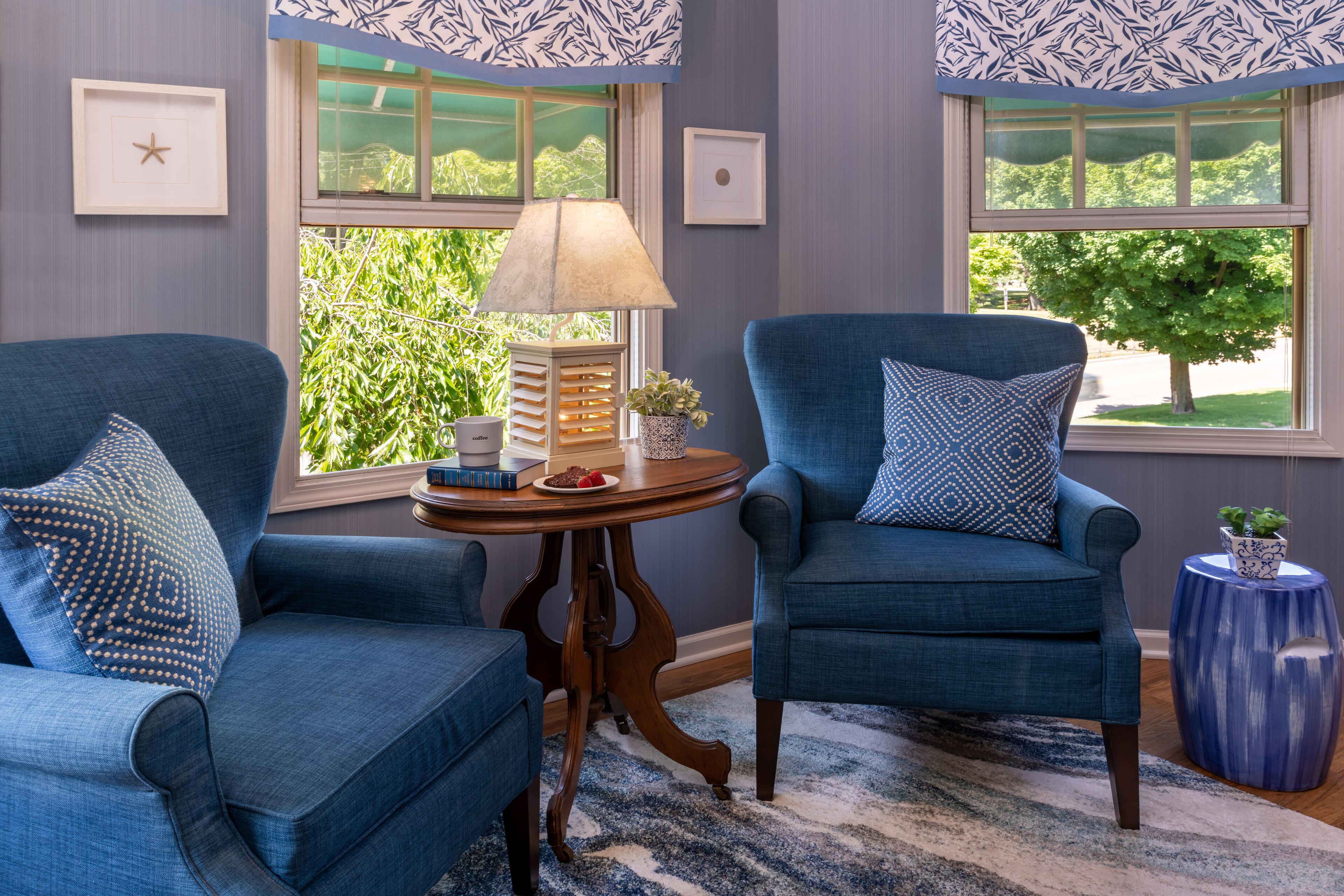 Une pièce avec deux fauteuils bleus, une petite table en bois surmontée d’une lampe, des tableaux encadrés accrochés au mur et une vue sur la verdure à l’extérieur.