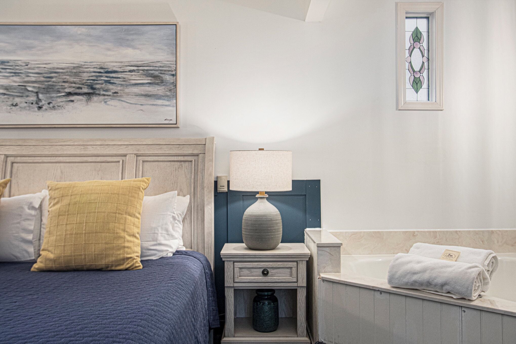 Une chambre bien rangée, avec un lit, une table de chevet, une lampe et une œuvre d’art encadrée accrochée au mur.