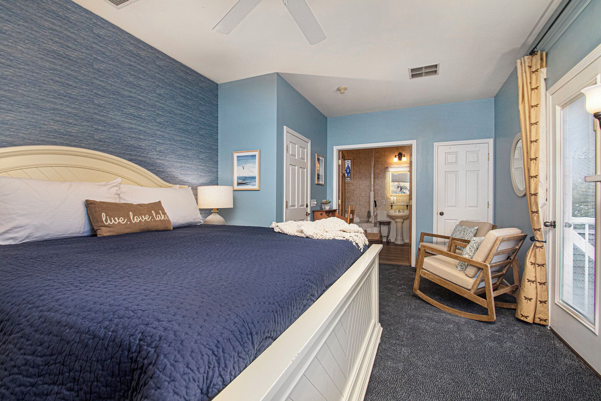 Une chambre à coucher avec un grand lit, un fauteuil à bascule et une salle de bain visible à travers une porte ouverte.
