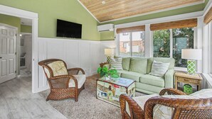 Una sala de estar con pared verde, techo de madera, muebles de mimbre, un sofá verde y un televisor montado en la pared.