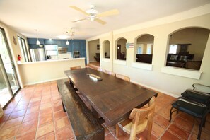 A spacious dining area with a large wooden table and benches, a ceiling fan, and a kitchen in the background.