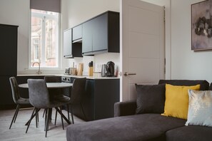 A modern kitchen with dark cabinets, a dining area with two chairs, and a sofa with a yellow pillow. - Apartment 3 (York)
