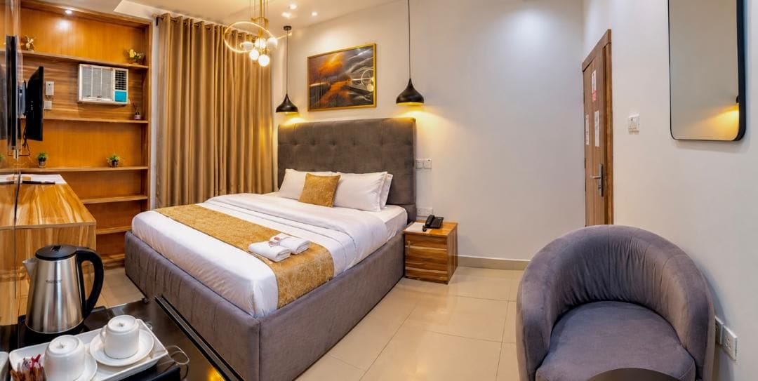 A hotel room with a bed, bedside table, chair, and a desk with a coffee maker and cups.