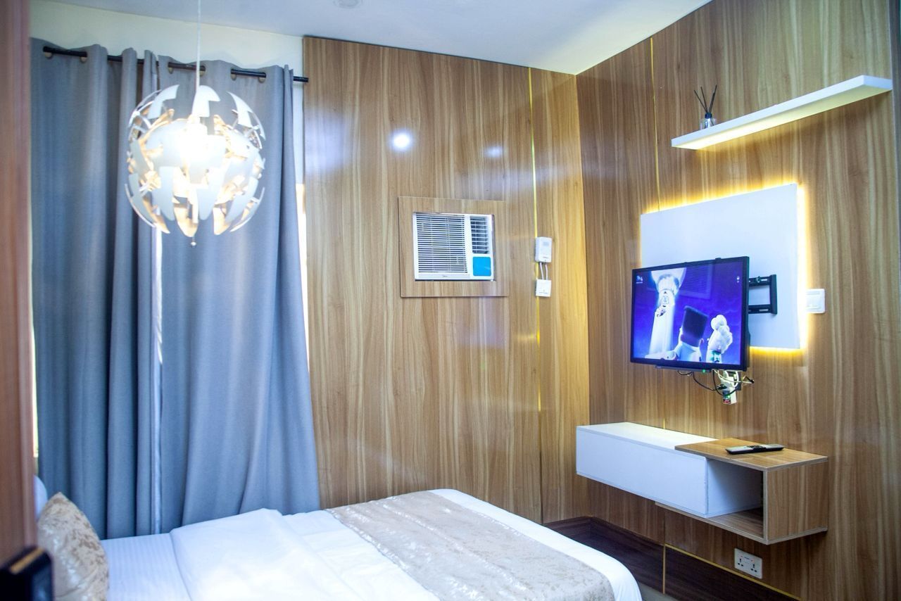 A modern hotel room with a wooden panel wall, a large bed with a white headboard, a wall-mounted TV, and a built-in shelf.