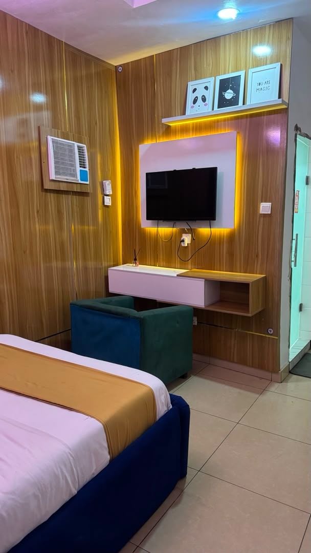 A hotel room with a bed, a TV mounted on a wooden wall, a desk with a chair, and a wardrobe.