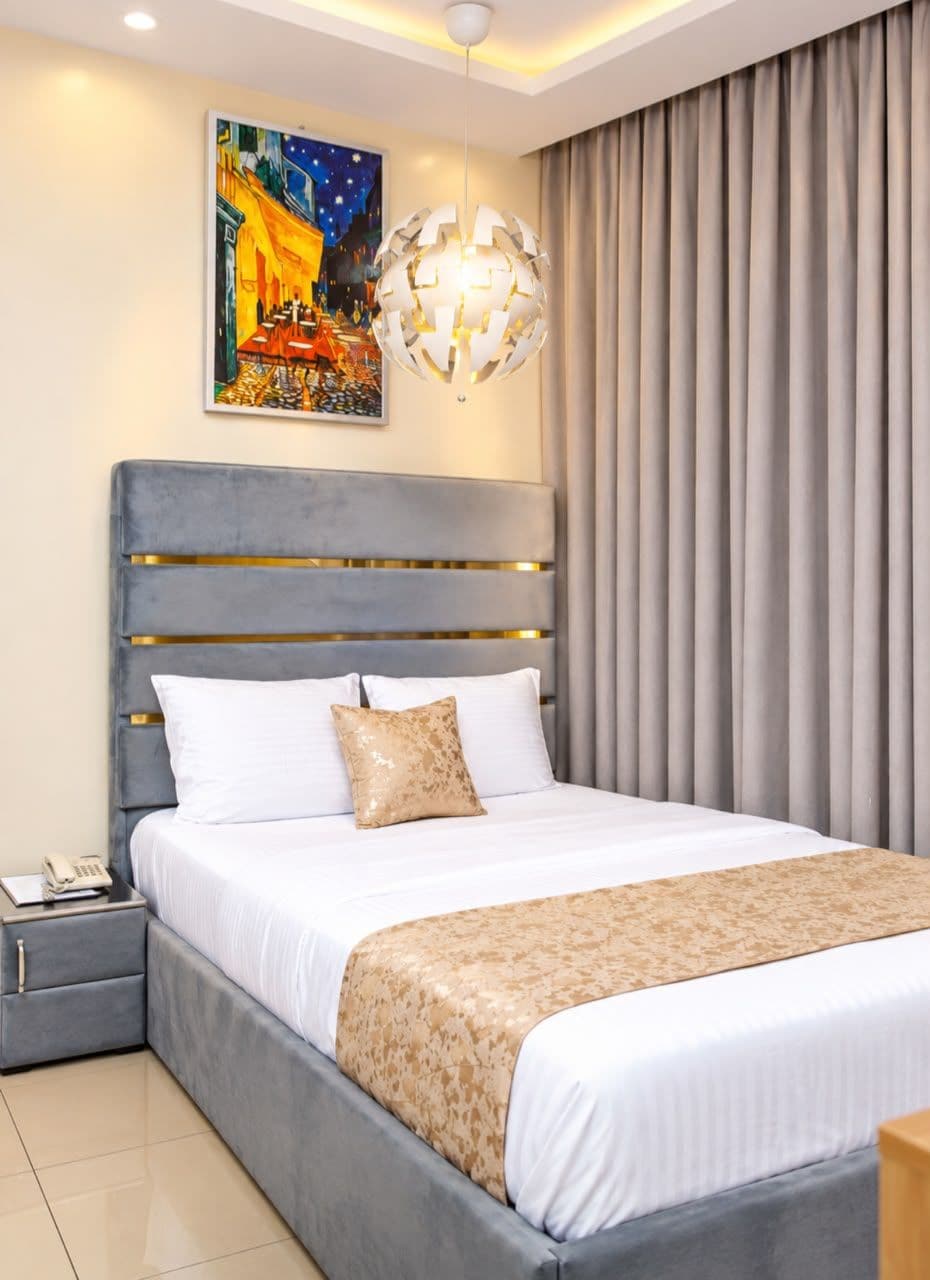 A modern hotel room with a large bed, a bedside table with a lamp, a painting on the wall, and a window with curtains.
