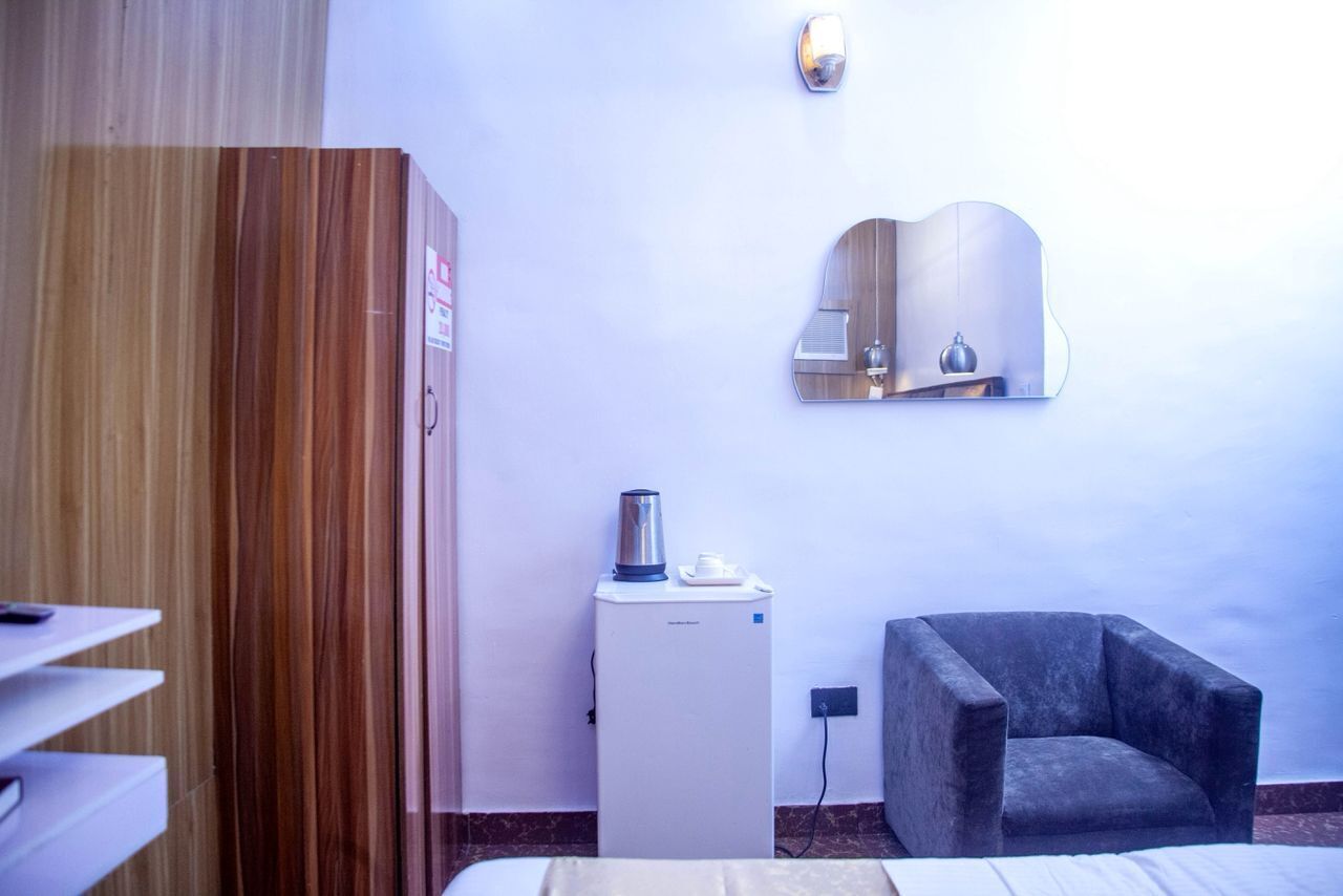 A hotel room with a wooden wardrobe, a small refrigerator, a chair, and a wall-mounted mirror.