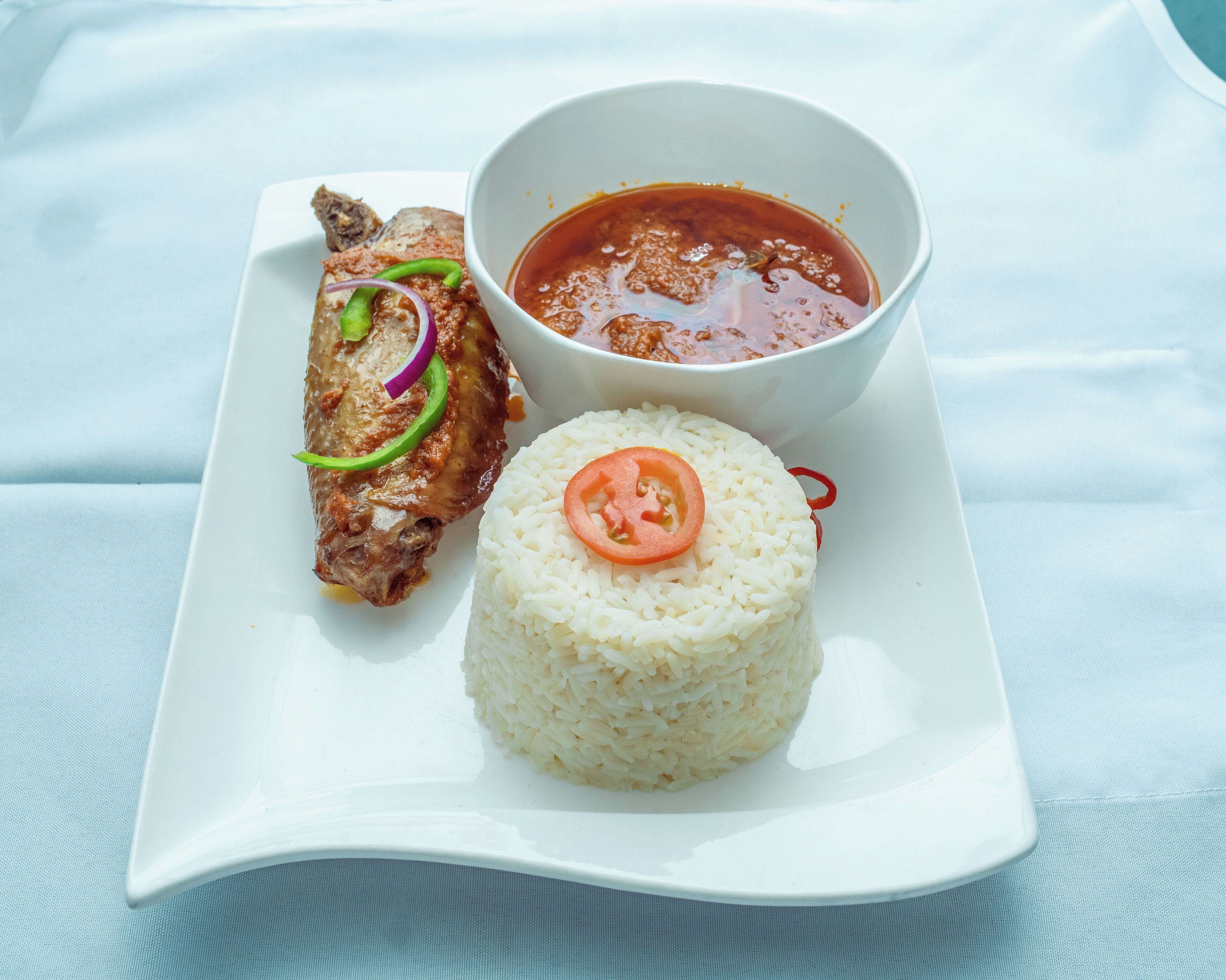 Une assiette de riz blanc garnie d’une tranche de tomate, d’un morceau de viande agrémenté de verdure et de rouge, et un bol de sauce rouge.