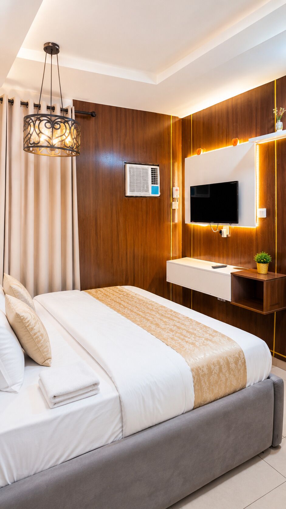 Ein modernes Hotelzimmer mit Bett, Fernseher, Schminktisch mit Spiegel und Deckenleuchte.