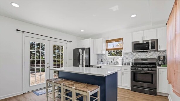 A modern kitchen with white cabinets, a central island with stools, stainless steel appliances, and a view through a glass door. - Beachfront 4BR - 30ft to Sand •Near Amusement Park (Keansburg)