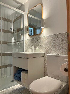 Un baño moderno con cabina de ducha de vidrio, un lavabo blanco con salpicadero de mármol y un inodoro con un rollo de papel higiénico.