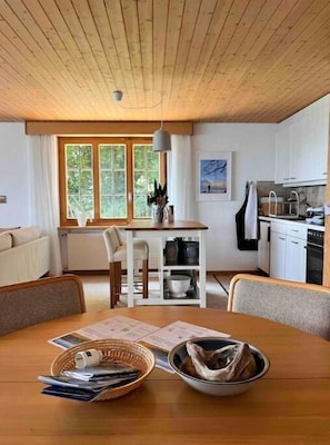 A cozy kitchen with a wooden table, a dining area, and a view of the outdoors through large windows.