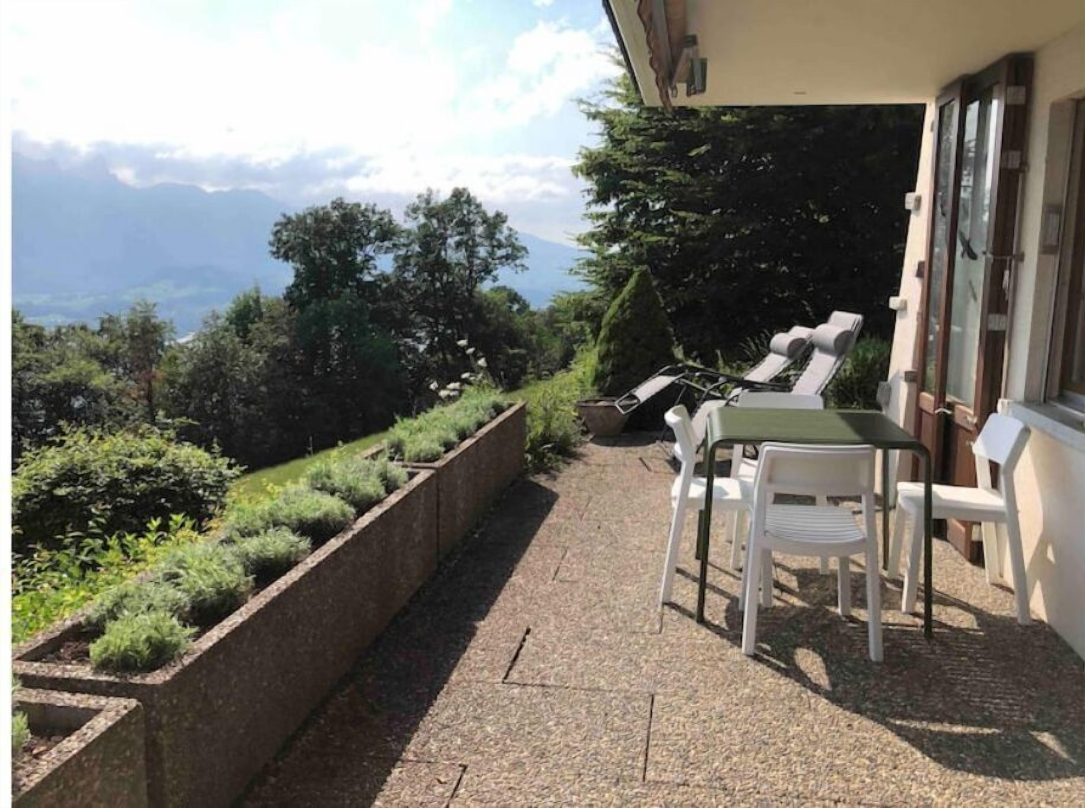 A balcony with a view of mountains, a table with chairs, and a garden bed with plants.