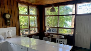 Une cuisine avec un plan de travail blanc, des armoires en bois, une horloge et de grandes fenêtres donnant sur des arbres.