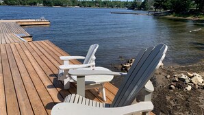 Une terrasse en bois avec des fauteuils Adirondack et une petite table, le tout situé au bord d’un lac.