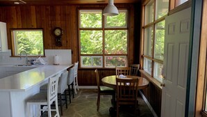 Une cuisine dotée d’un grand îlot central, de tabourets de bar blancs, d’une table à manger avec des chaises, d’une horloge et de grandes fenêtres offrant une vue sur la verdure.