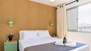 A hotel room with a bed, two nightstands with lamps, a window with a view, and a bottle of champagne on the bed.