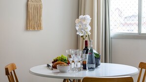 A round table with a white macrame wall hanging, a white orchid, a bottle of BOSS, a glass of water, a bowl of cereal, a loaf of bread, and a glass of wine.