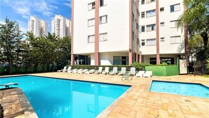 A swimming pool with lounge chairs, surrounded by a landscaped area with trees and a building in the background.