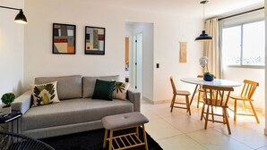 A modern living room with a grey sofa, a small round table, and wooden chairs. There are two abstract paintings on the wall and a potted plant on the sofa.