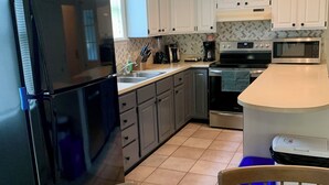 Eine moderne Küche mit weißen Schränken, einem schwarzen Kühlschrank, einer Mikrowelle und einer Kaffeemaschine. Es gibt einen Esstisch mit Stühlen und eine blaue Mülltonne.