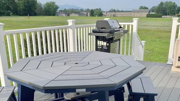 Eine Terrasse mit rundem Tisch und Bänken, umgeben von einem weißen Geländer, mit Blick auf eine Rasenfläche.