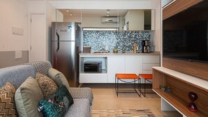 A compact living space with a kitchenette, a sofa with patterned cushions, and a flat-screen TV. - Condomínio Tag Decor (São Paulo)